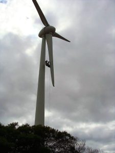 A wind turbine stands tall against a cloudy sky, with technicians suspended by ropes performing maintenance on one of its blades. Bushes are visible at the base of the turbine.