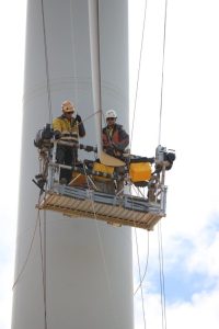 Two workers wearing safety gear are suspended on a platform high above the ground, performing maintenance on a large wind turbine. They are surrounded by harnesses and ropes against a backdrop of a cloudy sky.