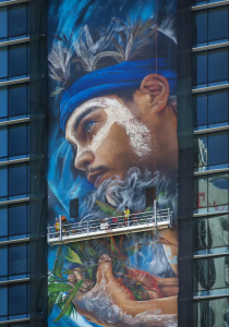 A large, vibrant mural depicting a person with a blue headband and face paint, holding leaves, is being painted on the side of a building. Workers on a scaffold are adding details to the artwork, which spans several floors.